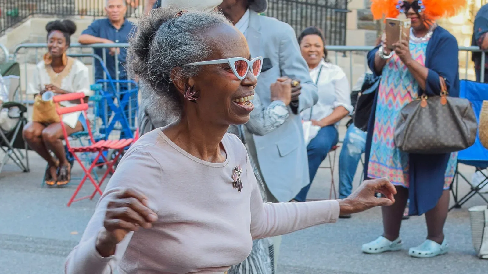 A joyful older Black woman with grey hair and white sunglasses dancing at a street party