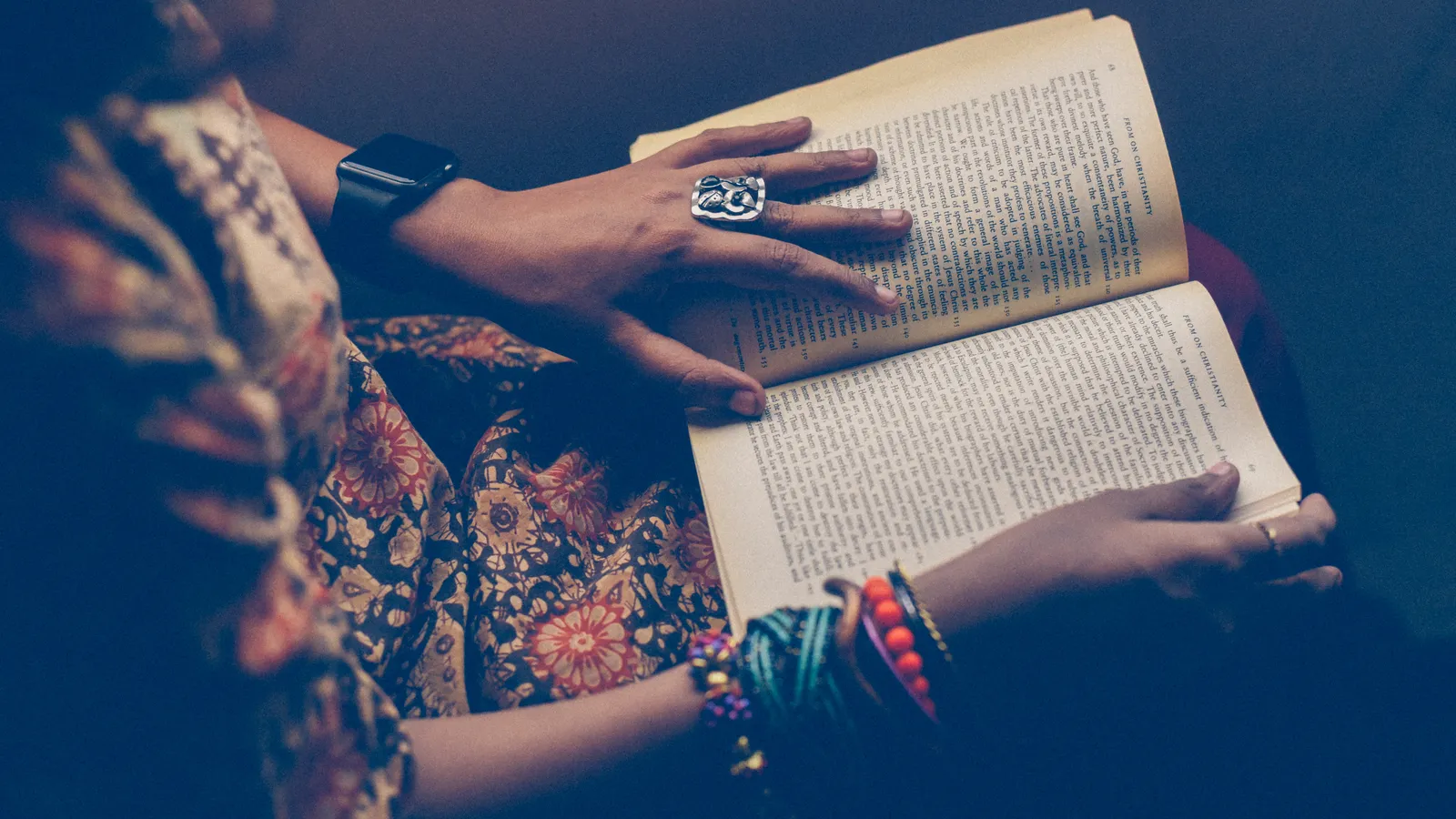 A woman in a patterned top and beaded bracelets reading an open paperback on her lap