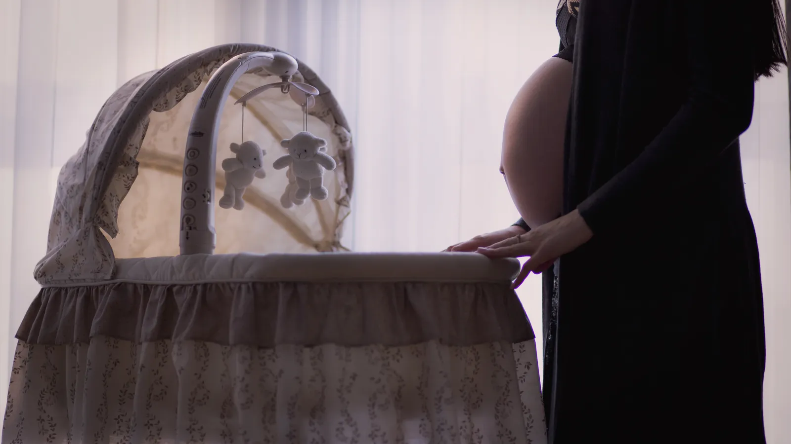 A pregnant woman standing beside a baby's crib in soft window light