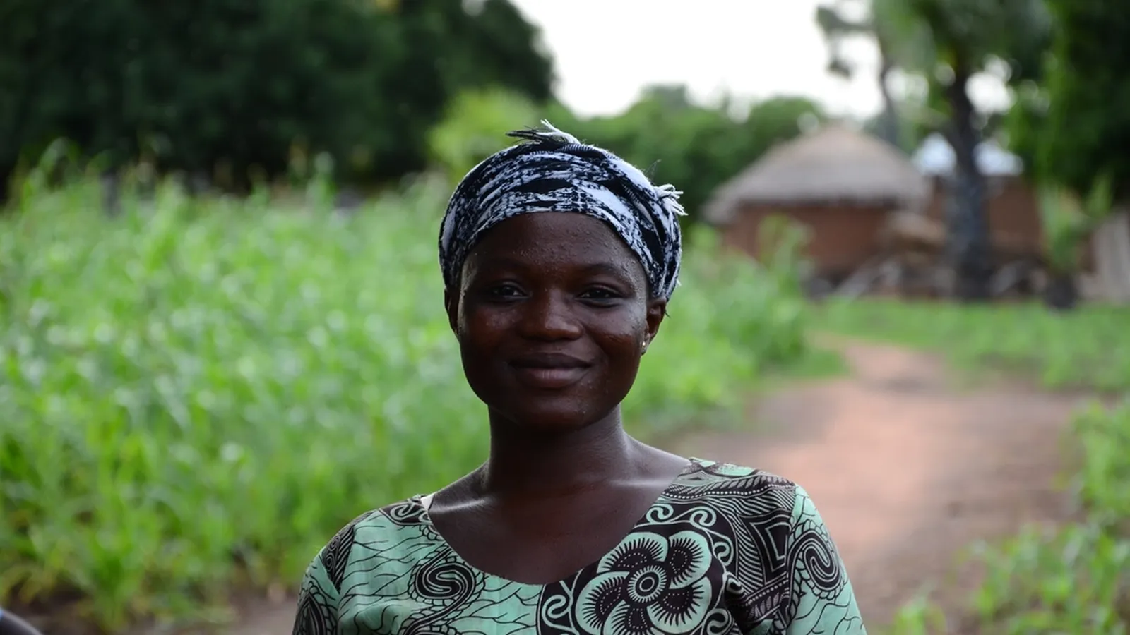 A smiling woman in a patterned headwrap standing outdoors on a warm green path, representing midlife women's health