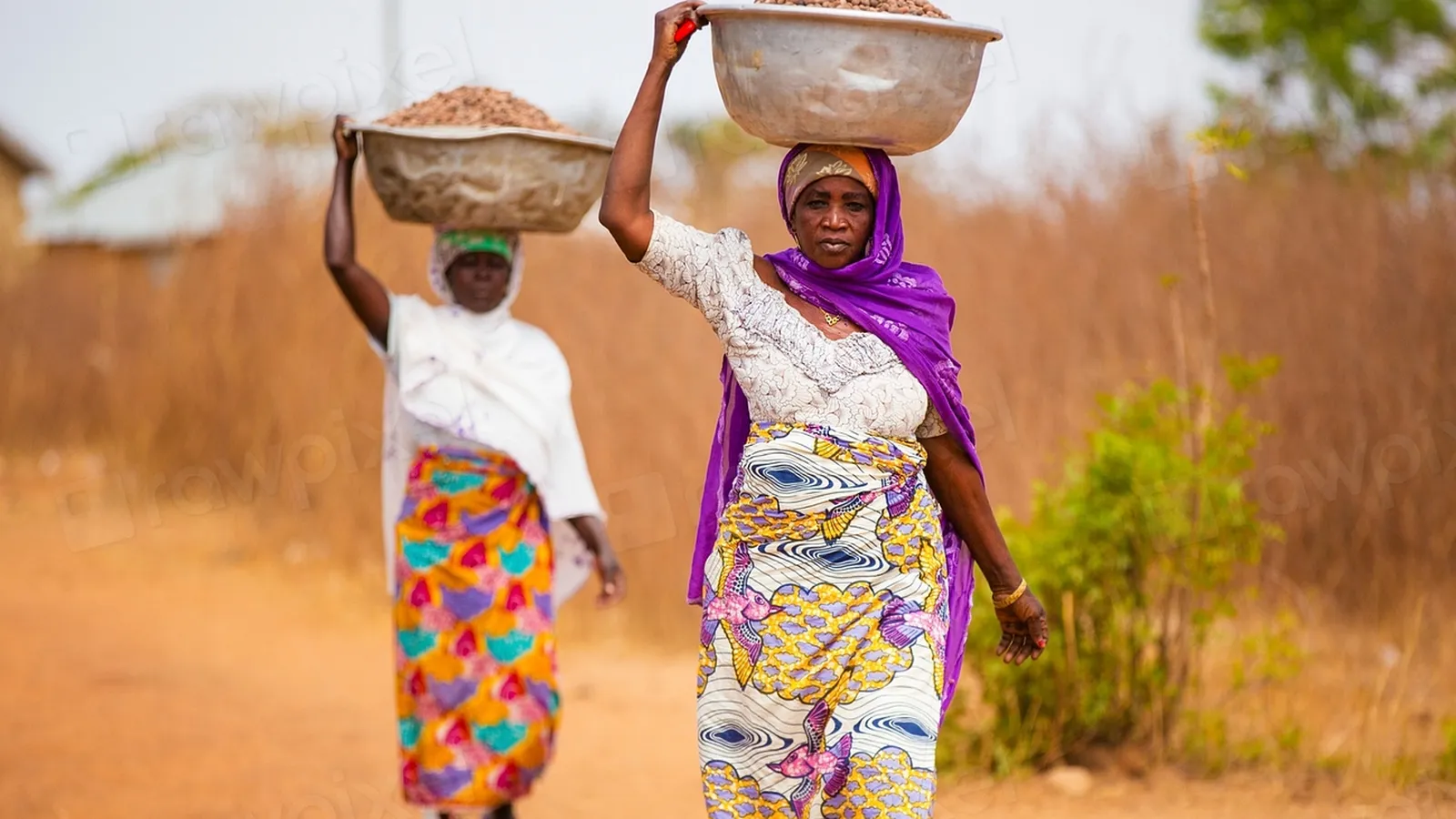 Two Ghanaian women in colourful wrappers walking along a dusty path carrying bowls on their heads