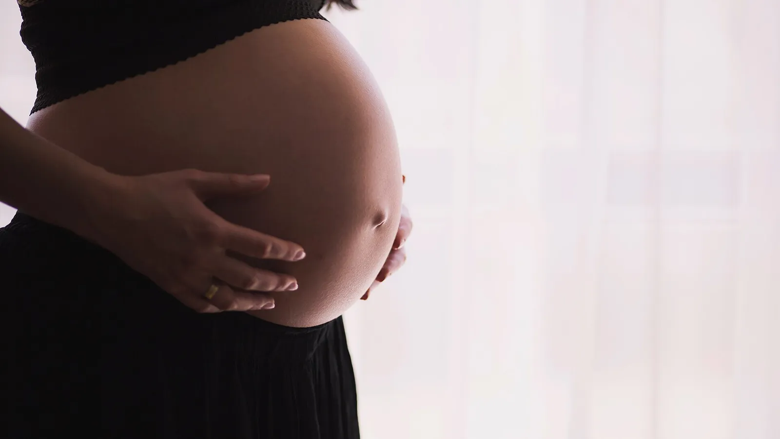 A pregnant woman in a dark top and skirt holding her bump against soft window light